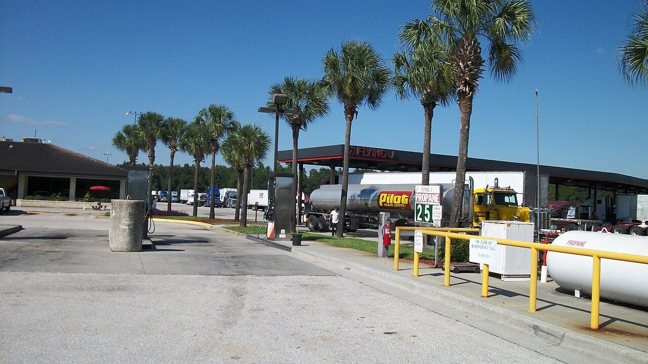A diesel tanker truck parked at a US truck stop with fuel pumps and palm trees